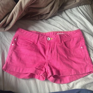 Rosa shorts från Adidas - Säljer ett par knallrosa shorts från Adidas med klassisk femficksdesign och knappgylf. De har bälteshällor och är perfekta för varma dagar. Adidas-loggan syns diskret på baksidan. Supersnygga till sommaren!