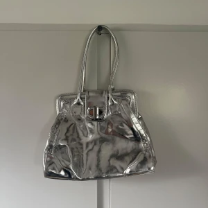 Silvrig axelväska  - Säljer en snygg och trendig axelväska i metallic silver. Väskan har ett stilrent spänne framtill och dubbla handtag. Använd men med mycket omsorg. 