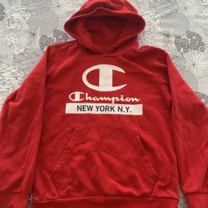 Röd hoodie från Champion med tryck - Säljer en röd hoodie från Champion med stor vit logga och texten 'NEW YORK N.Y.' på bröstet. Tröjan har huva och magficka, samt långa ärmar. Perfekt för dig som gillar streetwear och sportig stil. Ena ärmen har en pytte liten olje fläck men inget som syns mycket. 
