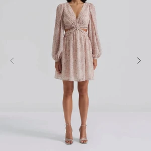 By Malina klänning med cut-out och ringdetalj - Supersöt ljusrosa kortklänning med småblommigt mönster, v-ringning och långärmade puffärmar. Klänningen har en snygg cut-out i midjan och en dekorativ ring framtill. Perfekt för vår och sommar! Helt ny med prislappen kvar, slutsåld överallt. Ny pris 3300kr. Skriv vid frågor!💕