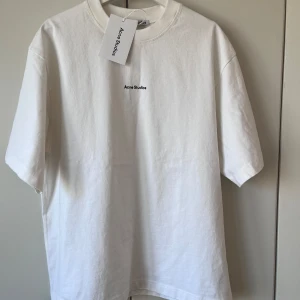 Vit t-shirt från Acne Studios - Vit t-shirt från Acne Studios med liten svart logga på bröstet. Klassisk rund halsringning och rak passform. Perfekt basic-plagg i mjuk bomull.