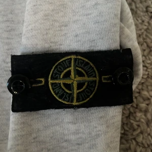 Ljusgrå sweatshirt från Stone Island - Säljer en ljusgrå sweatshirt från Stone Island med den klassiska svarta logotypen med gul brodyr på ärmen. Tröjan har rund hals och är tillverkad i mjuk bomull. Perfekt för en avslappnad och stilren look.