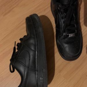 Svarta Nike Air Force 1 sneakers - Klassiska helsvarta Nike Air Force 1 sneakers med snörning och rund tå. De är lite slitna i hälen (se bilder) och det ena skosnöret har tappat plaständen. Skriv privat vid frågor eller fler bilder🤗