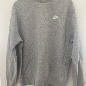 Grå sweatshirt från Nike - Klassisk grå sweatshirt från Nike med broderad vit logga på bröstet. Tröjan har rund halsringning och långa ärmar. Perfekt för en avslappnad och sportig stil.