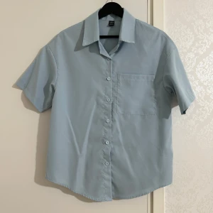 Ljusblå kortärmad skjorta från SHEIN - Ljusblå skjorta från SHEIN med korta ärmar och klassisk krage. Skjortan har knäppning framtill och en bröstficka. Perfekt för en stilren och enkel look.