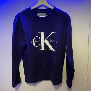 Mörkblå långärmad tröja från Calvin Klein Jeans - Säljer en mörkblå långärmad tröja från Calvin Klein Jeans med stor vit CK-logga framtill. Tröjan har rund halsringning och är i mjukt material, perfekt för en stilren och enkel look. Nypris ca 1000 kr mitt pris 300 kr