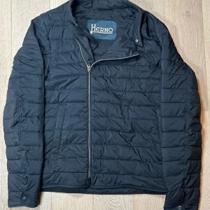 Svart quiltad jacka från Herno - Säljer en svart quiltad jacka från Herno med sned dragkedja framtill. Jackan är lätt vadderad och har lång ärm, perfekt för kyliga dagar. Enkel och stilren design som passar till mycket.