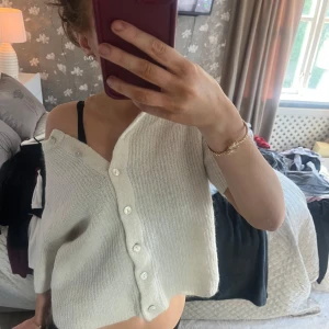 Vit croppad kofta med knappar - Säljer en vit, croppad kofta med knappar framtill. Tröjan har korta ärmar och är stickad i ett mjukt material. Perfekt för lager-på-lager och enkel att styla till olika outfits.