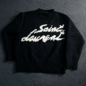 Saint Laurent stickad tröja  - Säljer en svart stickad tröja från Saint Laurent med vit, fluffig logotyp framtill. Tröjan har rund halsringning, långa ärmar och breda ribbade muddar vid ärmslut och nederkant. Perfekt för dig som gillar statement-plagg.☺️