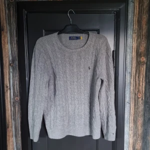 Grå kabelstickad tröja från Polo Ralph Lauren - Säljer en klassisk grå kabelstickad tröja från Polo Ralph Lauren i storlek M för att jag har växt hur den(Jag är 190 cm och väger 78 kg). Tröjan har rund halsringning, lång ärm och den ikoniska broderade loggan på bröstet. Perfekt för lager-på-lager och passar till det mesta. Påsen kommer med.