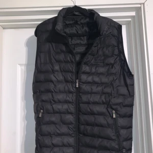 Svart dunväst från BOSS - Säljer en svart quiltad dunväst från BOSS med dragkedja framtill och två fickor med dragkedja. Västen har en hög krage och stilren design, perfekt för lager-på-lager under kyliga dagar.