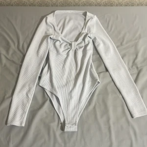 Vit ribbad body med rosett - Säljer en vit ribbad body med lång ärm och djup v-ringning framtill. Framtill finns en söt rosettdetalj. Perfekt att styla med jeans eller kjol för en trendig look.