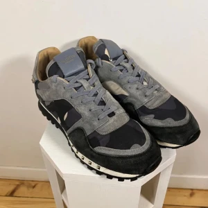 Valentino Rockrunner sneakers i grå och svart - Snygga Valentino Rockrunner sneakers i grått och svart med kamouflagemönster. Skorna har snörning, rund tå och detaljer i mocka och skinn. Perfekta för dig som vill ha en stilren men ändå unik look. En tag saknar å kan gå ner till 900 minst