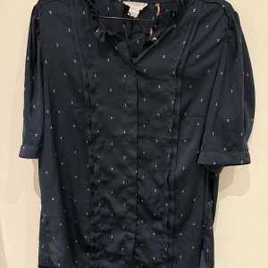 Mörkblå blus med ankarmönster - Säljer en mörkblå blus från Maison Scotch med små vita ankare som mönster. Blusen har korta ärmar och knappar framtill. Materialet känns svalt och lätt, perfekt för varmare dagar. Snygg och stilren med en maritim touch.