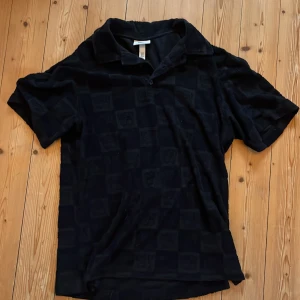 Svart t-shirt med krage från H&M x Smiley - Svart t-shirt med krage från H&M x Smiley. T-shirten har korta ärmar och ett diskret rutmönster i tyget. Perfekt för en avslappnad och stilren look. Sitter bra för storlek S och M också.