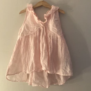 Ljusrosa blus - Säljer ett ljusrosa linne med volangdetaljer runt halsringningen och axlarna. Toppen är ärmlös och har en lös, flowy passform med lätt skrynkligt tyg. Perfekt för varma dagar och somriga outfits.