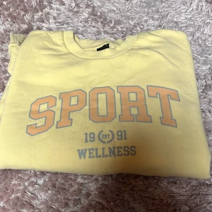 Sweatshirt från Gina  - Säljer en ljusgul sweatshirt med trycket 'SPORT 1991 WELLNESS' framtill. Tröjan har rund halsringning och långa ärmar. Den är köpt från Gina och är i storlek Medium.