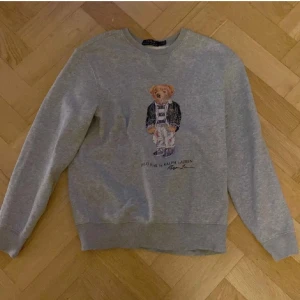 Grå sweatshirt med Polo Bear från Ralph Lauren - Säljer en grå sweatshirt från Ralph Lauren med det klassiska Polo Bear-motivet framtill. Tröjan har rund halsringning och långa ärmar. Perfekt för en avslappnad och stilren look.