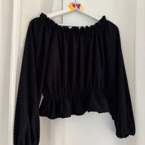 Svart off-shoulder tröja med broderade prickar - Svart blus med lång ärm och resår vid ärmslut och midja. Blusen har volang nertill och en lätt prickig struktur i tyget.🖤