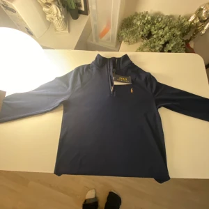 Mörkblå långärmad tröja från Polo Ralph Lauren - Säljer en mörkblå långärmad tröja från Polo Ralph Lauren med halv dragkedja och klassisk broderad logga på bröstet. Perfekt för en stilren och avslappnad look. Hög krage och mjukt material.