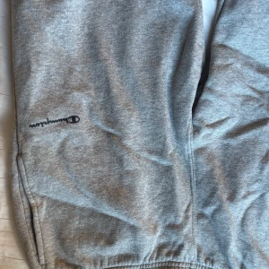 Grå mjukisbyxor från Champion - Grå mjukisbyxor från Champion med broderad logga på benet. Byxorna har resår i midjan och vid bensluten samt en dragkedjeficka på sidan. Perfekta för chill eller träning.