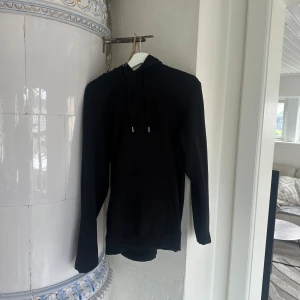 Svart hoodie från Les Deux - Säljer en svart hoodie från Les Deux med broderad logga på bröstet. Tröjan har huva med dragsko och en stor magficka. Perfekt för en avslappnad stil och enkel att matcha med det mesta.