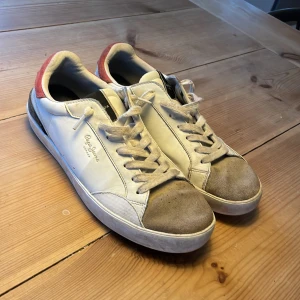 Vita sneakers från Pepe Jeans med röda detaljer - Säljer ett par vita sneakers från Pepe Jeans med röda detaljer på hälen och grå mocka framtill. Beat up design. Skorna har snörning, rund tå och platt sula. Perfekta för en avslappnad stil. Knappt använda.