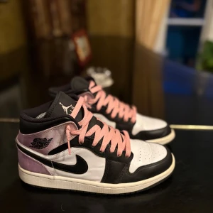 Nike Air Jordans med rosa detaljer - Säljer ett par Nike Air Jordans med svart och vit bas samt lila detaljer på hälen. Skorna har rosa snörning och klassisk Jordan-logga på sidan. Perfekta för dig som vill sticka ut med snygga sneakers.