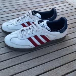 Adidas Sambas skor  - Säljer ett par klassiska Adidas Samba sneakers i vitt skinn med röda ränder och mörkblå detaljer. Skorna har platt sula, rund tå och snörning. Perfekta för dig som gillar stil! Storlek 45 1/3. Använda, fast i riktigt fint skick! 