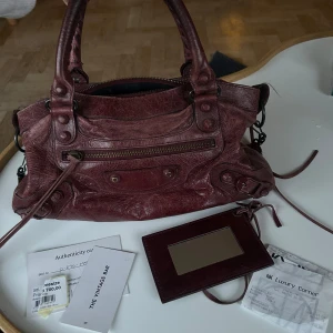 Vinröd handväska från Balenciaga - Säljer vinröd city bag i minsta storleken från Balenciaga i skinn med klassiska detaljer och dragkedja framtill. Väskan är köpt på The Vintage Bar på NK för 6700kr för ca 1,5 år sedan så äkthetsbevis och kvitto därifrån medföljer. Är i använt men fint skick. Har ett mindre hål på handtaget (se bild 3), se resterande bilder för skick i övrigt med exempelvis ”corner wear” och några slitningar på nitdetaljerna. Dock inget som påverkar väskans helhetsintryck. Pris kan diskuteras vid snabb affär 💕