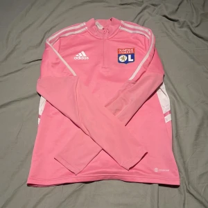 Rosa långärmad Olympique Lyonnais fotbollströja från Adidas - Säljer en rosa långärmad fotbollströja från Adidas med Olympique Lyonnais klubbmärke på bröstet. Tröjan har vita detaljer och dragkedja vid halsen. Perfekt för dig som vill sticka ut på planen eller träningen.
