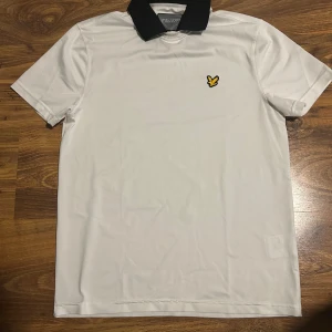 Vit piké från Lyle & Scott med svart krage - Säljer en stilren vit pikétröja från Lyle & Scott med svart krage och klassisk gul logga på bröstet. Tröjan är kortärmad och tillverkad i ett syntetmaterial som känns lätt och bekvämt.