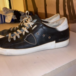 Svarta sneakers från Philippe Model - Svarta sneakers från Philippe Model med vita detaljer och vit sula. Skorna har snörning och en klassisk låg profil. Tillverkade i skinn med märkets logga på sidan och bak på hälen.