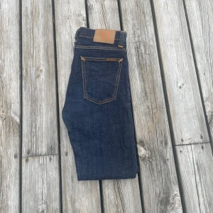 Mörkblå jeans från Nudie Jeans - Säljer ett par mörkblå jeans från Nudie Jeans med klassisk femficksdesign och kontrastsömmar. Byxorna har en rak passform och är tillverkade i denim med snygga detaljer på bakfickan. Perfekta för dig som gillar stilrena jeans! Modellen är high kai. Ny pris ligger på runt 1600kr!