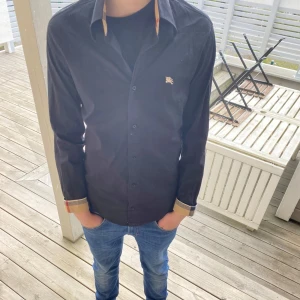 Svart skjorta från Burberry med klassiska detaljer - Säljer en svart skjorta från Burberry med klassiskt rutigt tyg på insidan av kragen och manschetterna. Skjortan har långa ärmar, knappar framtill och en liten broderad logga på bröstet. Perfekt för dig som gillar stilrena plagg med lyxig känsla.