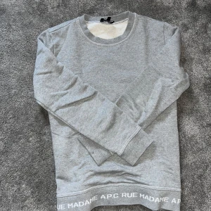 Grå sweatshirt från A.P.C. - Säljer en stilren grå sweatshirt från A.P.C. med texten 'RUE MADAME A.P.C.' längs nederkanten. Tröjan har rund halsringning och långa ärmar. Perfekt för en avslappnad och trendig look.
