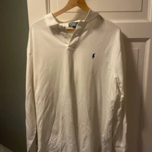 Vit långärmad pikétröja från Polo Ralph Lauren - Klassisk vit långärmad pikétröja från Polo Ralph Lauren med broderad logga på bröstet. Tröjan har krage och knäppning vid halsen, perfekt för en stilren och tidlös look.