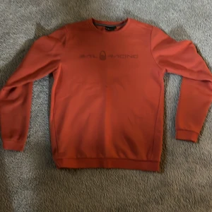 Orange sweatshirt från Sail Racing - Säljer en orange sweatshirt från Sail Racing med rund halsringning och diskret logga framtill. Tröjan har långa ärmar och ribbade muddar vid ärmslut och nederkant. Perfekt för en avslappnad stil.