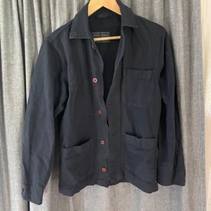 Mörkblå overshirt med röda knappar - Säljer en mörkblå overshirt med tre stora fickor framtill och röda knappar. Jackan har klassisk krage och lång ärm, perfekt för lager-på-lager. Enkel och stilren design som funkar till mycket. Köpt i arbetarboden i stockholm nästan helt oanvänd. Väldigt fin jacka som är tillverkad i italien av ett litet lokalt företag som gör allt förhand och tar beställningar. Org pris 2000. Skriv för frågor :)