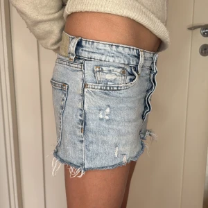 Ljusblå jeanskjol med råa kanter - Säljer en ljusblå jeanskjol med slitna detaljer och råa kanter. Kjolen har klassiska fickor fram och bak samt bälteshällor. Perfekt för dig som gillar en avslappnad och trendig stil.