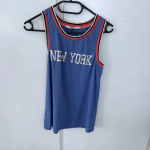 Blå ärmlös meshtröja med New York-tryck från ONLY - Säljer en blå ärmlös meshtröja från ONLY med vitt New York-tryck framtill. Tröjan har sportig känsla med röda och vita detaljer runt hals och ärmhål. Perfekt för en avslappnad och cool stil.