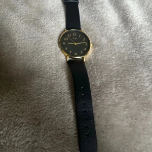 Svart och guldig analog klocka från Regal behövs byte på batteri  - Stilren analog klocka från Regal med svart läderband och rund urtavla i svart. Klockan har guldfärgade detaljer och siffror, vilket ger en klassisk look. Perfekt accessoar för att lyfta din outfit.
