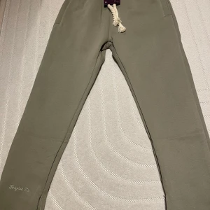 Syna world Tracksiut - Säljer ett par beiga mjukisbyxor och hoodie från Syna World med broderad logga på benet och coolt tryck baktill. Byxorna har dragsko i midjan med grovt snöre och elastisk midja för extra komfort.