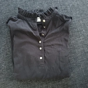 Svart blus med volangkrage och knappar - Säljer en svart blus från Holly&Whytte med volangdetalj vid kragen och knappar framtill. Perfekt för dig som gillar en stilren men ändå söt look. Långärmad modell med mjukt bomullsmaterial.