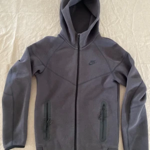 Mörkgrå Nike tech från Nike - Säljer en mörkgrå hoodiejacka från Nike med dragkedja framtill och två fickor med dragkedja. Jackan har en sportig look med huva och diskret Nike-logga på bröstet. Perfekt för dig som gillar stilren och enkel design.