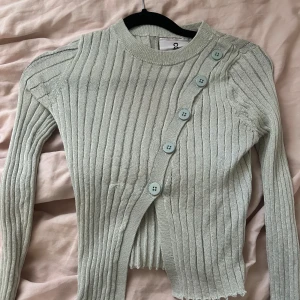 Ljusgrön ribbad tröja med sned knäppning - Säljer en ljusgrön ribbad tröja med lång ärm och sned knäppning framtill med stora knappar. Tröjan har en tight passform och är tillverkad i mjukt material. 