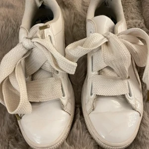Vita sneakers Puma Basket - Säljer ett par klassiska vita Puma sneakers med breda vita snören och guldlogga på sidan. Skorna har en låg profil, rund tå och platt sula. Perfekta för en stilren look
