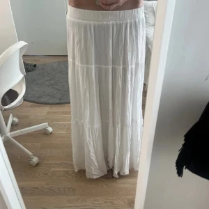 Vit långkjol med volang - Säljer en luftig och flowy vit långkjol med volangdetaljer. Kjolen har resår i midjan och är tillverkad i ett lätt material som ger ett snyggt fall. Perfekt för varma dagar och enkel att styla med olika toppar.