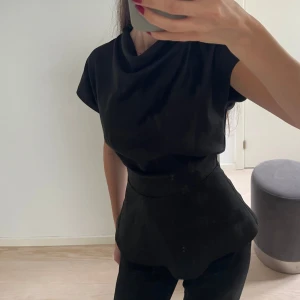 Svart satintopp med knytning i nacken - Säljer en svart satintopp med korta ärmar och snygg drapering framtill. Toppen har en öppen rygg med knytning i nacken som ger en elegant touch. Perfekt att styla med både jeans och kjol.