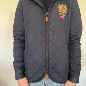 Morris jacka - Säljer en svart quiltad jacka från Morris med guldbroderad logga och brittisk flagga på bröstet. Jackan har dragkedja framtill och klassisk krage. Perfekt för dig som gillar stilren design och snygga detaljer.